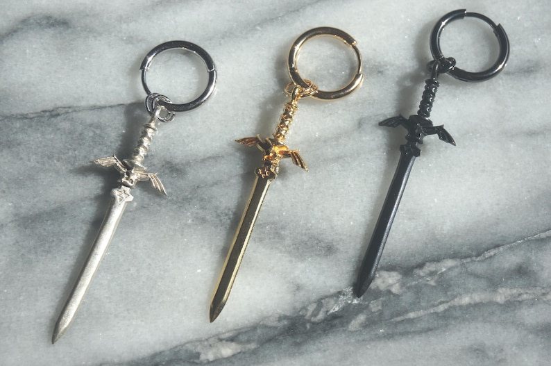 May include: Three sword-shaped earrings with hoop closures. The earrings are silver, gold, and black.