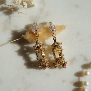 May include: Gold-coloured chandelier earrings with detailed designs. Each earring has a dangling, ornate structure with clear and pink accents. The earrings are presented on a marble surface, with a sprig of dried flowers and a strand of pearls.
