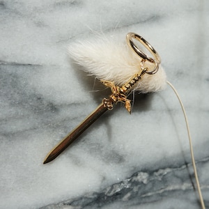 May include: A gold sword charm with a hoop for attaching to a necklace or bracelet. The charm is on a white fluffy background.