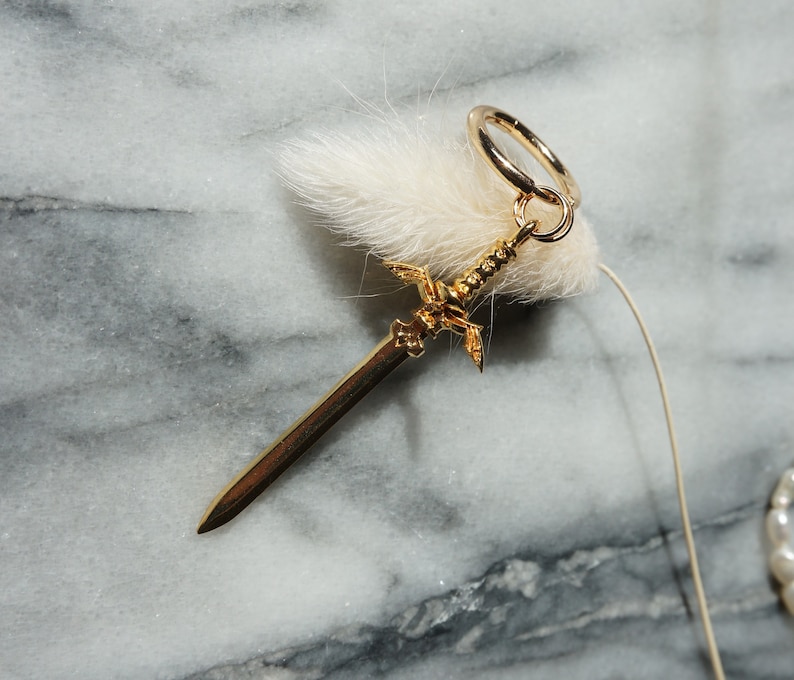 May include: A gold sword charm with a hoop for attaching to a necklace or bracelet. The charm is on a white fluffy background.