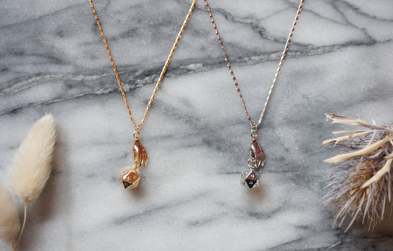 May include: Two gold necklaces with a hand holding a gold and silver dice pendant. The necklaces are on a marble surface.