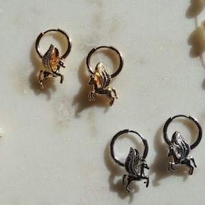 Puede incluir: Pendientes de aro dorados y plateados con un dije de Pegaso. Los pendientes se exhiben sobre una superficie de mármol, con un collar de perlas y flores secas en el fondo. El Pegaso tiene alas detalladas.