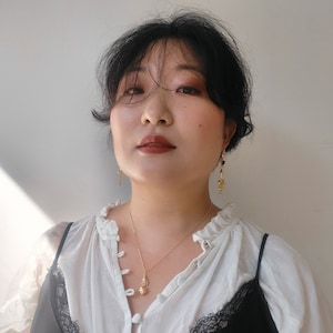 May include: A woman with dark hair wearing a white blouse with a ruffled collar and a gold necklace with a small pendant. She is also wearing gold earrings.