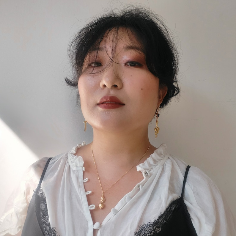 May include: A woman with dark hair wearing a white blouse with a ruffled collar and a gold necklace with a small pendant. She is also wearing gold earrings.