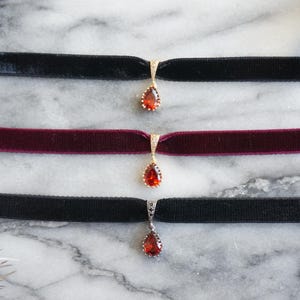 Puede incluir: Tres collares gargantilla de terciopelo con un colgante en forma de lágrima con una piedra roja. Las gargantillas son negras, burdeos y negras.