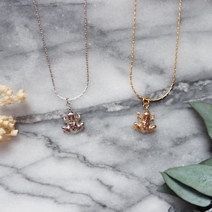 May include: Two necklaces featuring frog pendants. One is silver, the other gold. The frog pendants are detailed and three-dimensional. The chains are delicate, set against a marble surface.