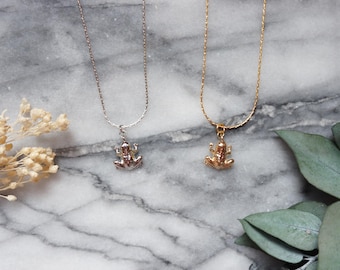 May include: Two necklaces with frog pendants. One necklace is silver, the other is gold. The frog pendants are detailed and three-dimensional. The chains are delicate and hang on a marble surface.