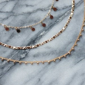 Pode incluir: Três colares de ouro sobrepostos em uma superfície de mármore. O colar superior tem pequenos pingentes redondos de ouro. O colar do meio é uma corrente grossa e texturizada. O colar inferior tem pequenas contas redondas de ouro.