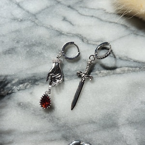 May include: A pair of silver-toned earrings. One earring features a hand pendant with a red teardrop-shaped gem. The other earring has a sword pendant. Both earrings have a hoop closure.