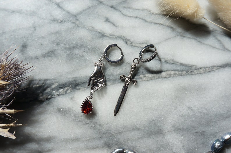May include: A pair of silver-toned earrings. One earring features a hand pendant with a red teardrop-shaped gem. The other earring has a sword pendant. Both earrings have a hoop closure.