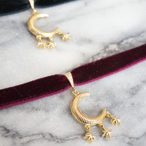 May include: Two velvet chokers with gold-coloured crescent moon pendants and star charms. One choker is black, the other is a deep burgundy. The background is a light grey and white marble.