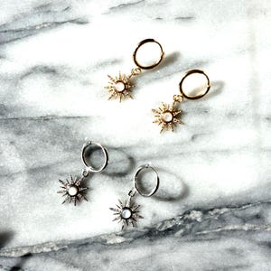 May include: Two pairs of earrings with a sunburst design. One pair is gold-toned, the other is silver-toned. Each earring features a small, round hoop and a sun-shaped pendant with a central pearl-like bead.