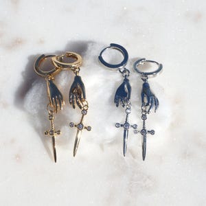 May include: Two pairs of hoop earrings, one gold-toned and one silver-toned. Each earring features a dangling charm of a hand holding a small sword. The sword charms are accented with tiny clear stones set along the blade and the crossguard.