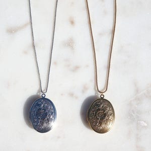 May include: Two oval-shaped lockets on thin metal chains. One locket is silver-toned and the other is gold-toned. Both lockets feature an intricate, embossed floral pattern on the front surface and hang from matching delicate link chains.