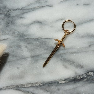 May include: A gold sword charm with wings, attached to a gold hoop earring.