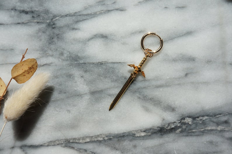 May include: A gold sword charm with wings, attached to a gold hoop earring.