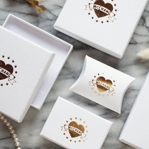 May include: White gift boxes with a heart and banner logo that reads "McChang". The logo is in brown and gold. One box is open, revealing a white interior. The boxes are arranged on a marble surface.