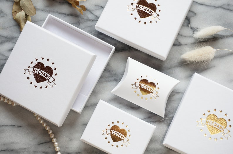 May include: White gift boxes with a heart and banner logo that reads "McChang". The logo is in brown and gold. One box is open, revealing a white interior. The boxes are arranged on a marble surface.