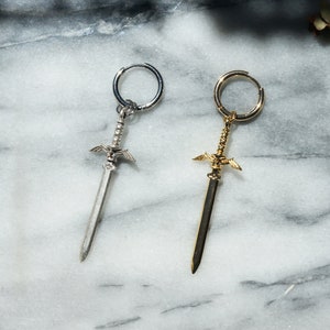May include: Two sword-shaped earrings, one silver and one gold, with a hoop design. The earrings are on a white marble surface.