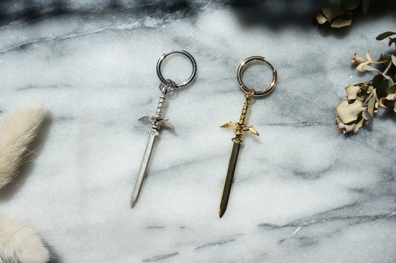May include: Two sword-shaped earrings, one silver and one gold, with a hoop design. The earrings are on a white marble surface.