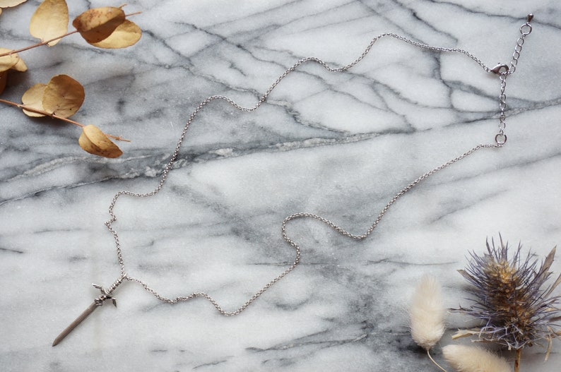 Pu&ograve; includere: Una collana color argento con un ciondolo a forma di spada. Il ciondolo ha un'elsa e una lama dettagliate. La catena &egrave; delicata e regolabile. La collana &egrave; esposta su una superficie di marmo con foglie secche.