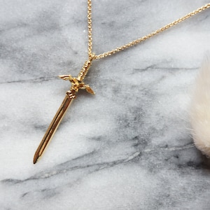 Pu&ograve; includere: Una collana con pendente color oro con un design a spada. La spada presenta dettagli intricati sull'elsa e sulla lama. La collana &egrave; esposta su una superficie di marmo, con una catena delicata.