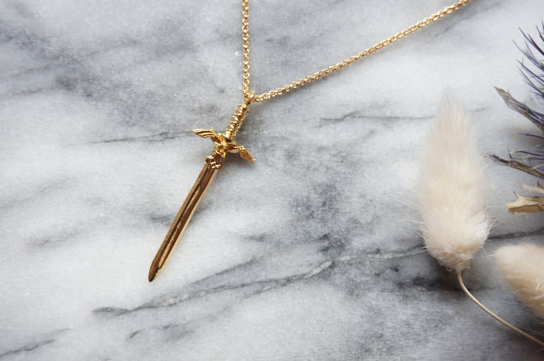 Pu&ograve; includere: Una collana con pendente color oro con un design a spada. La spada presenta dettagli intricati sull'elsa e sulla lama. La collana &egrave; esposta su una superficie di marmo, con una catena delicata.