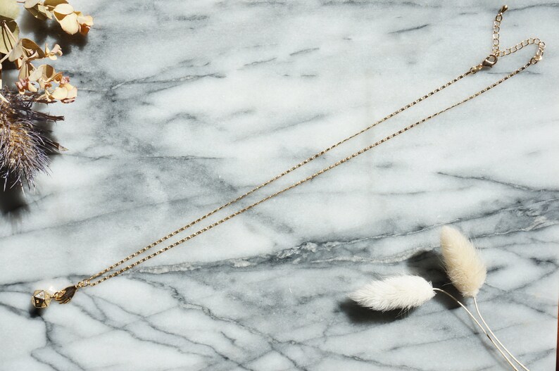 May include: A delicate gold chain necklace with a small, pearlescent pendant. The necklace is displayed on a marble surface with dried flowers.