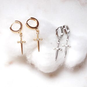 May include: Two pairs of hoop earrings featuring a dangling sword-shaped charm. One pair is gold-toned and the other is silver-toned. Each sword charm is accented with small, clear, round-cut stones set along the blade and the crossguard.