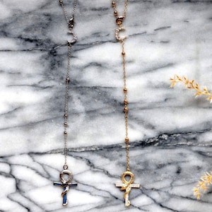 May include: Two Y-shaped necklaces, one silver and one gold, are shown against a marble backdrop. The silver necklace has a crescent moon pendant and an Ankh cross. The gold necklace features a similar design with a gold Ankh cross.