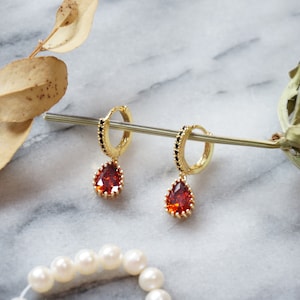 Puede incluir: Un par de pendientes de aro dorados con una piedra preciosa roja en forma de lágrima que cuelga. Los aros están acentuados con cuentas negras.