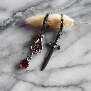 May include: A pair of gothic-style earrings. One earring features a hand pendant with a red teardrop-shaped gem. The other earring has a small sword pendant. Both earrings have black accents and are displayed on a marble surface.