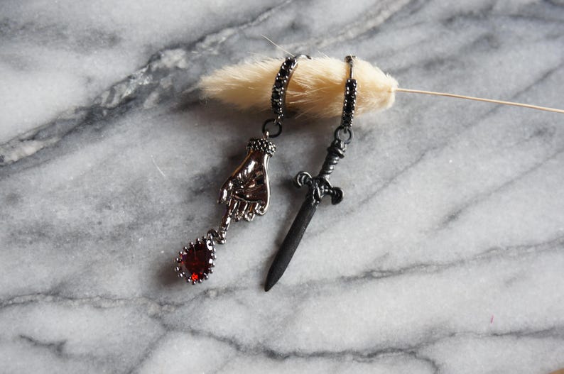 May include: A pair of gothic-style earrings. One earring features a hand pendant with a red teardrop-shaped gem. The other earring has a small sword pendant. Both earrings have black accents and are displayed on a marble surface.