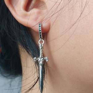 May include: Silver-toned dangle earring featuring a sword pendant. The earring has a small hoop with black stones and a detailed sword charm. The sword has a textured handle and a pointed blade, creating a gothic or fantasy aesthetic.