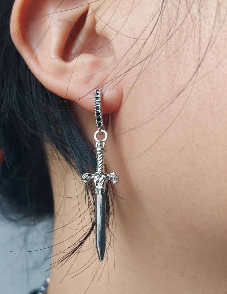 May include: Silver-toned dangle earring featuring a sword pendant. The earring has a small hoop with black stones and a detailed sword charm. The sword has a textured handle and a pointed blade, creating a gothic or fantasy aesthetic.