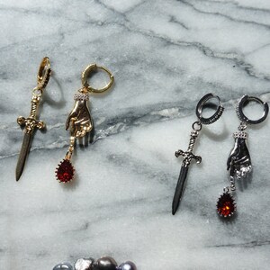 May include: Two pairs of dangle earrings. One pair is gold-toned with a sword and a hand holding a red teardrop gem. The other pair is silver-toned with a sword and a hand holding a red teardrop gem. The earrings are on a marble surface.