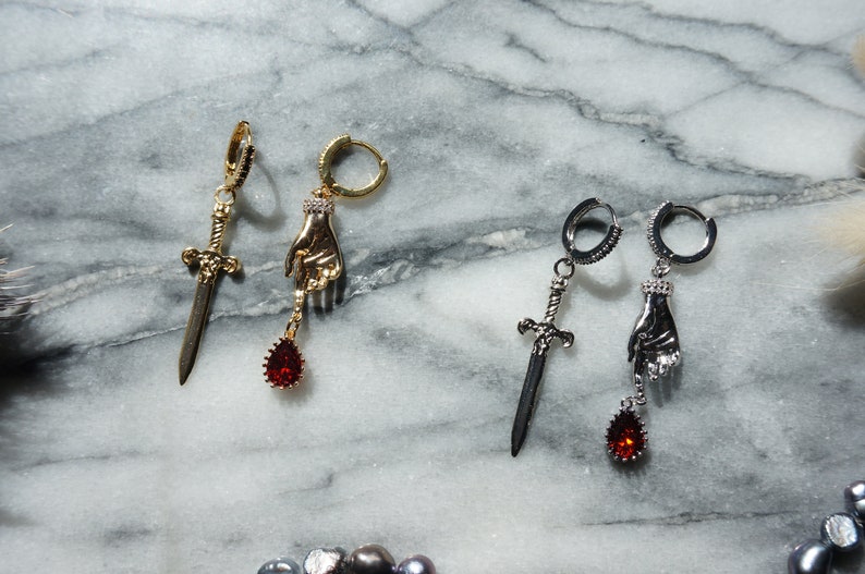 May include: Two pairs of dangle earrings. One pair is gold-toned with a sword and a hand holding a red teardrop gem. The other pair is silver-toned with a sword and a hand holding a red teardrop gem. The earrings are on a marble surface.