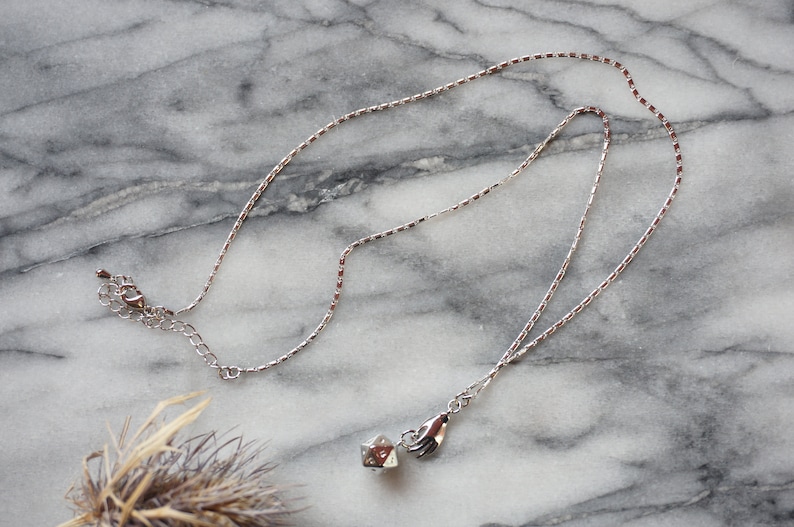 May include: A silver chain necklace with a small silver charm hanging from it. The charm is a cube with a hand holding a star on top.