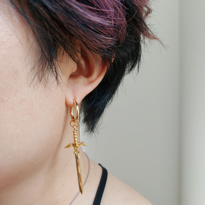 May include: Gold hoop earring with a sword charm dangling from it. The earring is worn in the left ear of a person with short, dark purple hair.