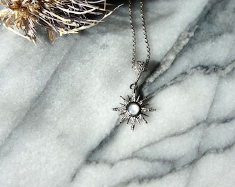 May include: A silver-toned sunburst pendant necklace with a central white stone and surrounding clear crystals. The pendant hangs from a delicate chain. The necklace is displayed on a marble surface, with a dried plant in the background.