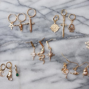 May include: A collection of gold-toned hoop and drop earrings featuring various charms including a star, a skull, a sword, a circular medallion, a hamsa hand, a snake, a starburst, a crescent moon, a hand, a green teardrop, and a beetle.