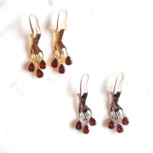 Peut inclure: Deux paires de boucles d'oreilles pendantes avec des breloques en forme de main métallique. Une paire est dorée et l'autre argentée. Chaque main tient trois cristaux rouges en forme de poire, facettés et aux bords métalliques dentelés.