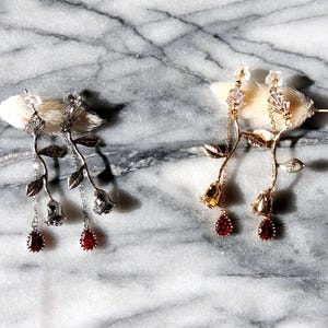 May include: Two pairs of elegant earrings. One pair is silver with red teardrop gems and rose details. The other pair is gold with red teardrop gems and rose details. Both pairs are displayed on a white surface against a marble background.