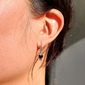 May include: A silver hoop earring with a black heart-shaped pendant. The earring has a row of small black stones along the hoop. The heart pendant is a solid black colour.