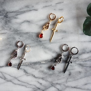 May include: An assortment of earrings with unique designs. The earrings feature a hand holding a red gem, and a sword. The earrings are gold, silver, and black. The background is a marble surface.