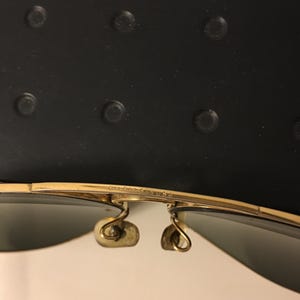 May include: Close-up of gold-rimmed sunglasses with dark lenses. The sunglasses have a classic aviator style with a double bridge and curved temples. The image shows the details of the frame and hinges.