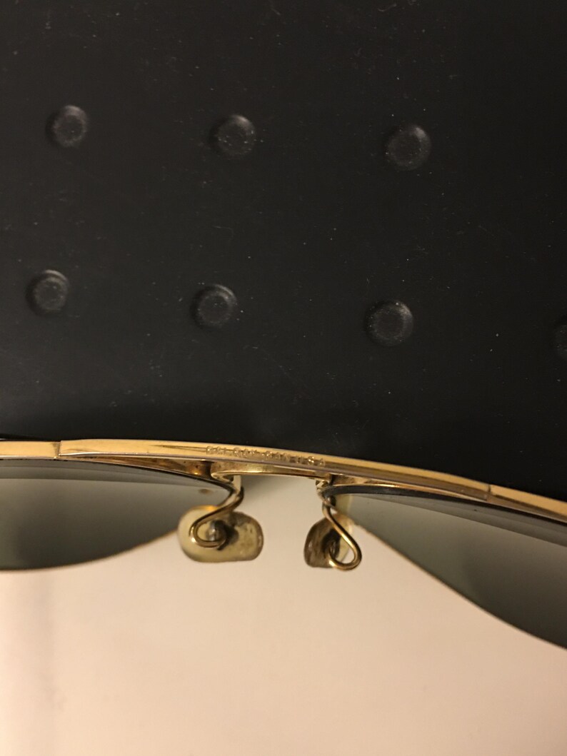 May include: Close-up of gold-rimmed sunglasses with dark lenses. The sunglasses have a classic aviator style with a double bridge and curved temples. The image shows the details of the frame and hinges.
