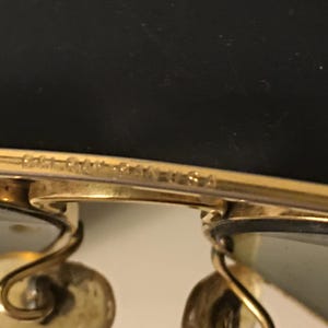 May include: Close-up of gold-toned sunglasses with a classic design. The frame has a delicate, curved shape, and the lenses are a light green color. The text "Bausch & Lomb USA" is visible on the frame.