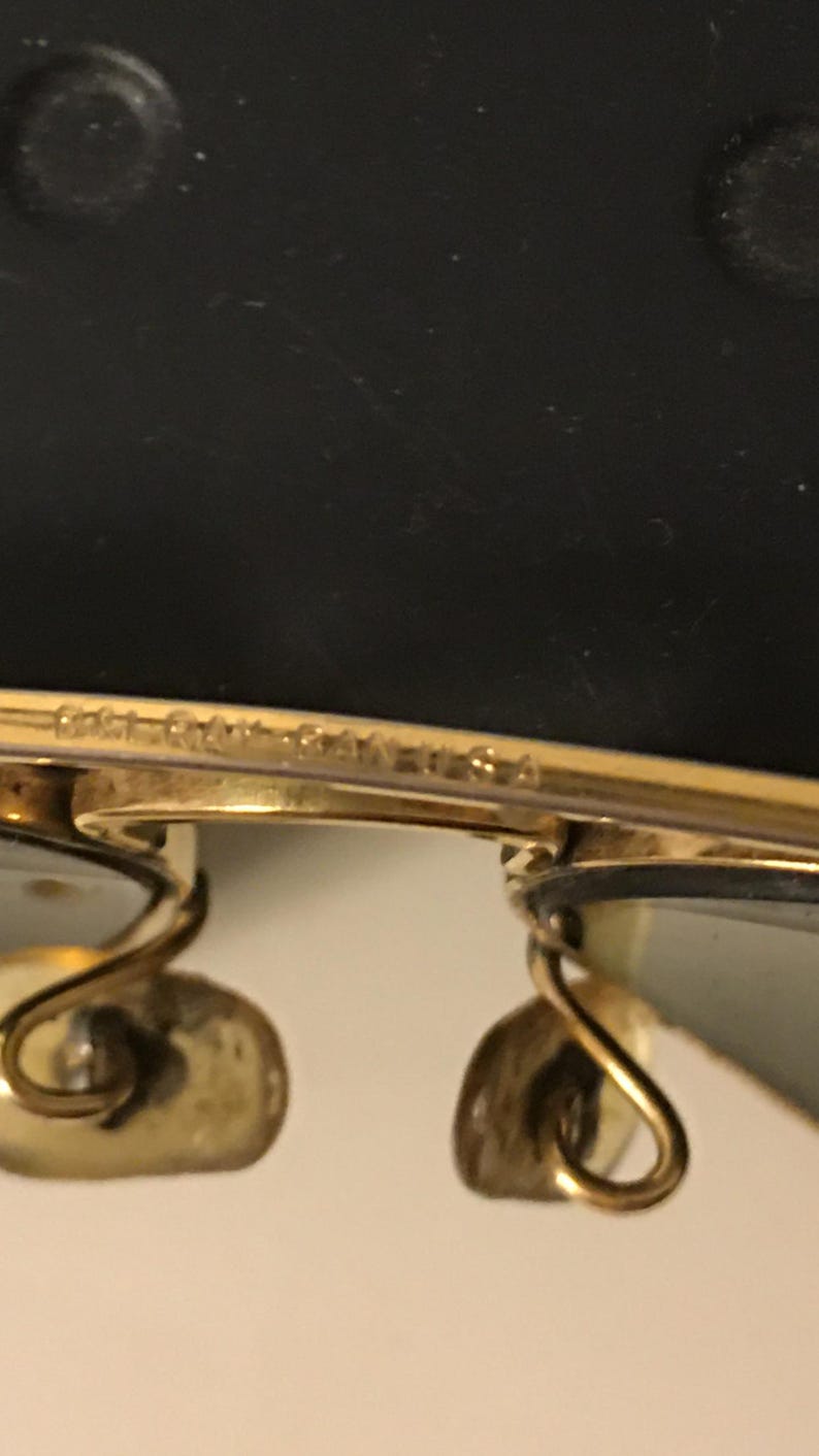 May include: Close-up of gold-toned sunglasses with a classic design. The frame has a delicate, curved shape, and the lenses are a light green color. The text "Bausch & Lomb USA" is visible on the frame.