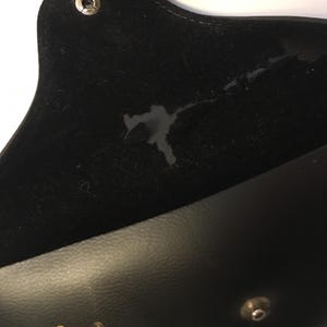 May include: Black eyeglass case with a snap closure. The interior is lined with black velvet, showing a stain. The exterior is smooth black leather. The case is open, revealing the interior.